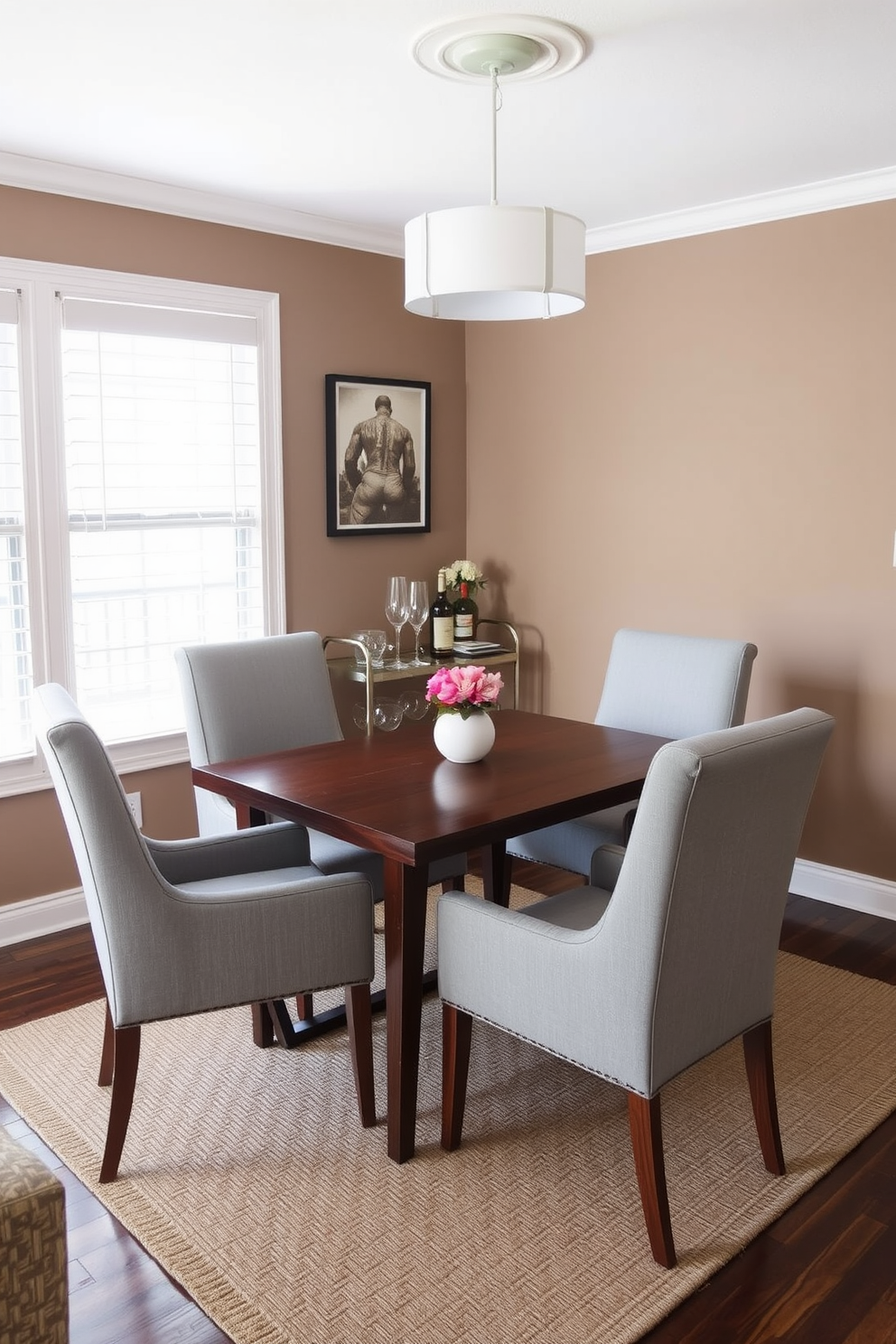 A cozy dining room featuring a small wooden table surrounded by four upholstered chairs in a soft gray fabric. In the corner, a stylish bar cart holds an assortment of glassware and a selection of spirits, complemented by a small vase of fresh flowers. The walls are painted in a warm taupe color, creating an inviting atmosphere. A modern pendant light hangs above the table, casting a soft glow over the space, while a textured area rug anchors the seating area.