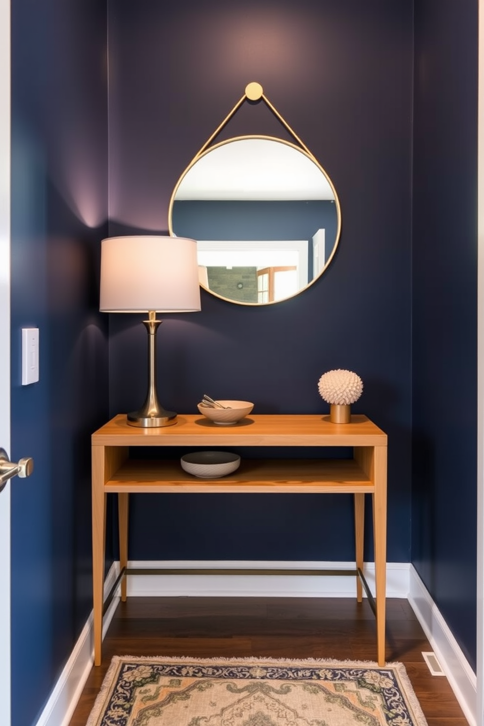 A bold accent wall painted in deep navy blue serves as the focal point of a small entryway. Complementing the wall, a sleek console table in natural wood is placed against it, adorned with a stylish lamp and a decorative bowl for keys. The entryway features a round mirror with a thin gold frame above the console table, enhancing the space's brightness. A small, patterned rug lies beneath the table, adding warmth and texture to the compact area.