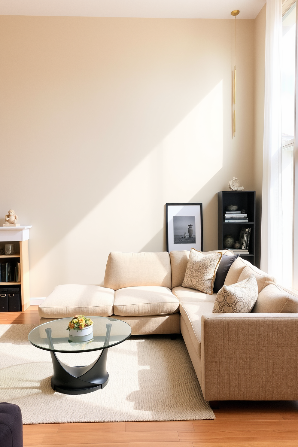 A compact sectional sofa is placed in a cozy living room with warm neutral tones. The walls are painted a soft beige, and a round coffee table with a glass top sits in front of the sofa. To the side, a stylish bookshelf filled with decorative items adds personality to the space. A large window allows natural light to flood in, complemented by sheer white curtains.