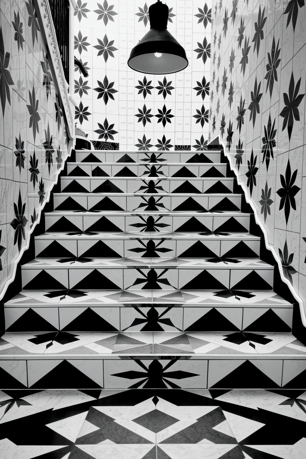 A striking monochrome patterned tile staircase features bold geometric designs in black and white. The staircase is illuminated by a modern pendant light that casts interesting shadows on the intricate tile work.