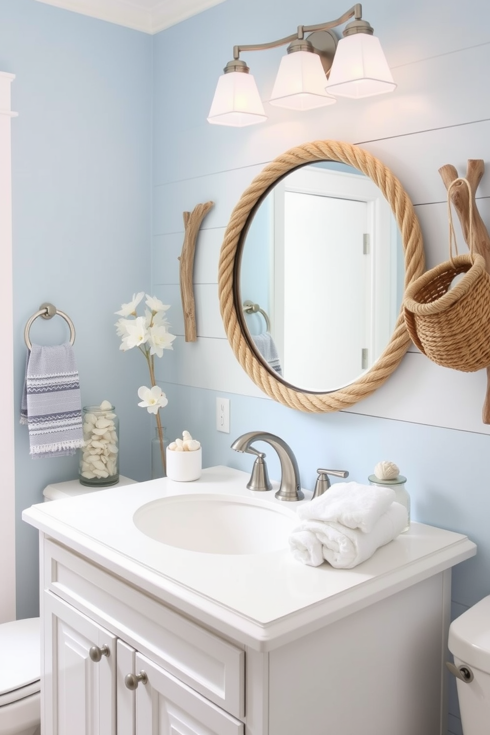 A serene summer bathroom setting inspired by nautical themes. The walls are painted in a soft blue hue, complemented by white shiplap accents and driftwood decor elements. A round mirror with a rope frame hangs above a white vanity with brushed nickel fixtures. Decorative items include seashells in a glass jar and a woven basket filled with fluffy white towels.