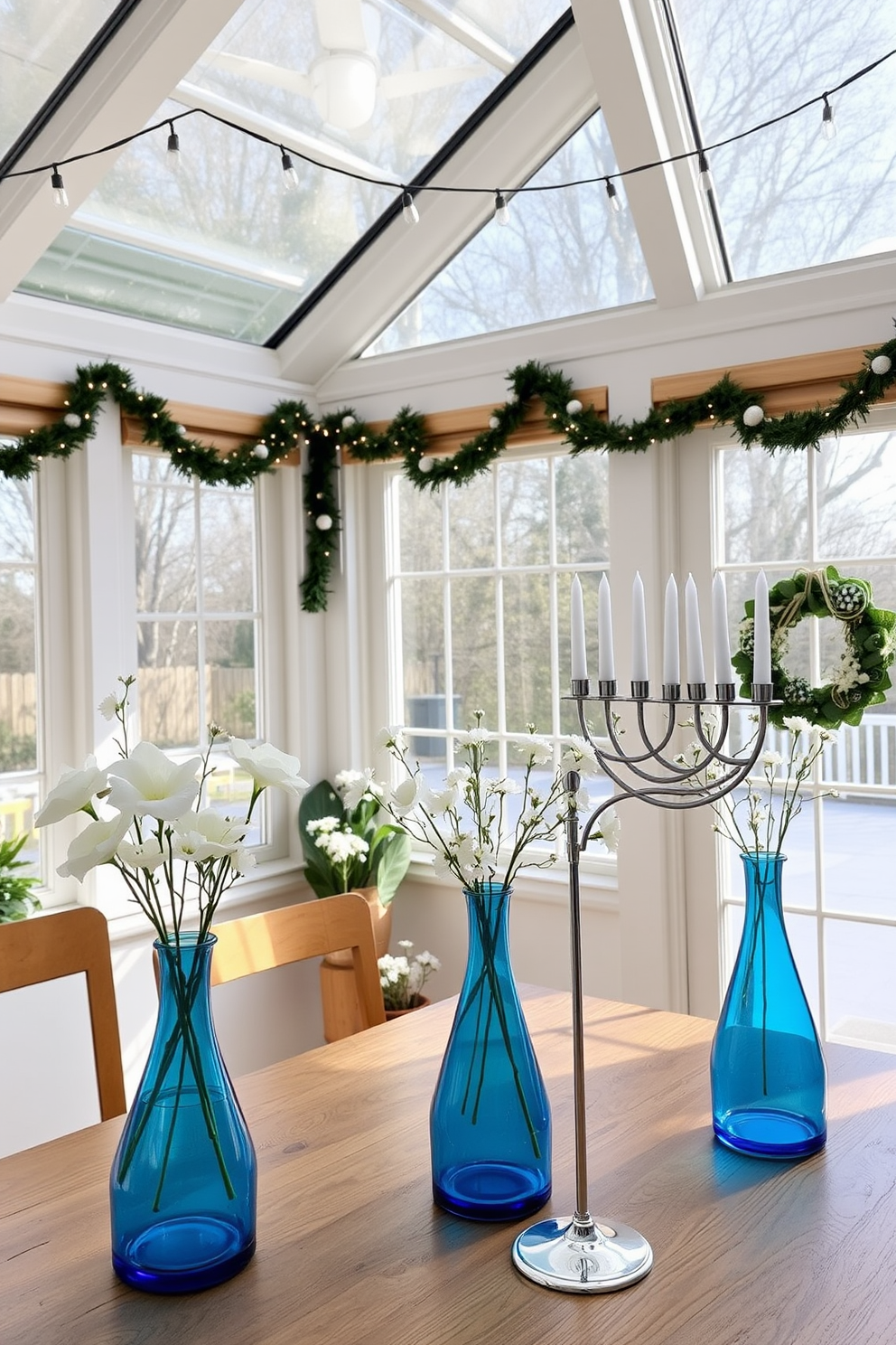 A bright sunroom filled with natural light. There are blue glass vases elegantly arranged on a wooden table, each holding fresh white flowers. Festive decorations for Hanukkah adorn the space. String lights twinkle above, and a beautifully designed menorah sits prominently on the table, complementing the serene atmosphere.