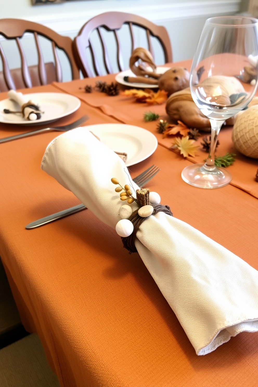 Handmade napkin rings crafted from natural materials adorn the elegantly set dining table. The table is draped with a warm, earthy tablecloth, and surrounded by rustic wooden chairs, creating a cozy atmosphere for Thanksgiving.