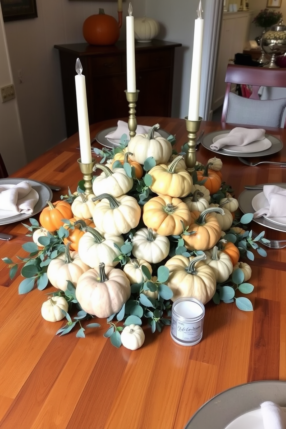 A charming table centerpiece featuring an assortment of mini pumpkins in various sizes and colors. Surrounding the pumpkins are eucalyptus leaves and small white candles to create a warm and inviting atmosphere for Thanksgiving. The table is set in a cozy small space, with a rustic wooden table that complements the autumn theme. Simple place settings with neutral-colored plates and linen napkins enhance the festive yet understated decor.