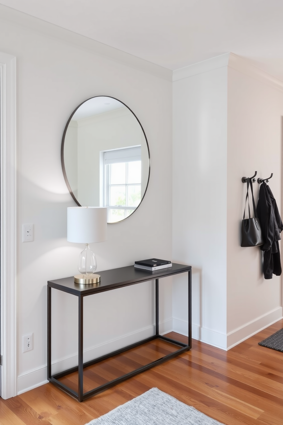 A stylish entryway features a sleek console table against the wall with a modern lamp on top. Above the table, a large round mirror reflects the natural light coming from the nearby window. On one side, there are stylish hooks mounted on the wall for hanging coats and bags. The flooring is a warm hardwood that complements the neutral color palette of the walls.