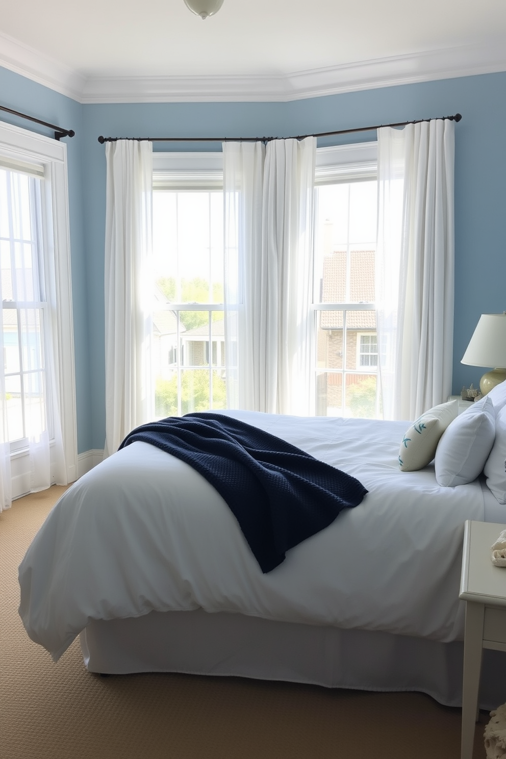 A serene townhouse bedroom with a coastal theme. The walls are painted in soft blue, and natural light floods in through large windows adorned with sheer white curtains. The bed is dressed in crisp white linens with a textured navy throw blanket. Decorative elements include seashells and driftwood accents, creating a relaxed beachy atmosphere.