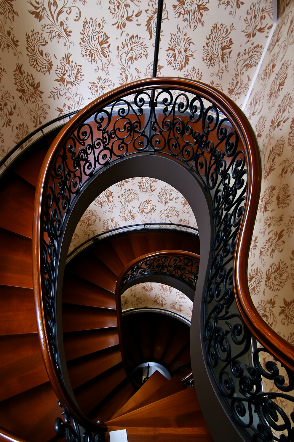 A stunning spiral staircase with intricate wrought iron railings and a polished wooden banister. The steps are made of rich mahogany, and the surrounding walls are adorned with vintage wallpaper featuring a floral pattern.