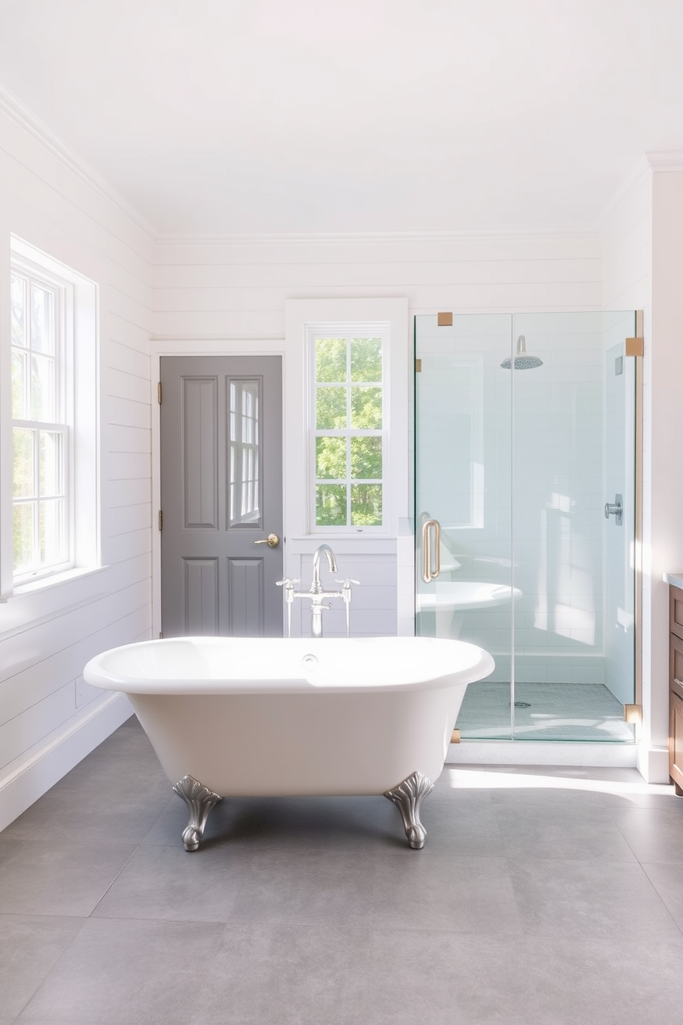 A classic clawfoot tub is elegantly positioned in the center of a bright and airy bathroom. The walls are adorned with soft white shiplap, and large windows allow natural light to flood the space. A sleek glass shower enclosure stands adjacent to the tub, featuring brushed nickel fixtures. The floor is covered with large gray tiles, providing a contemporary contrast to the vintage charm of the clawfoot tub.