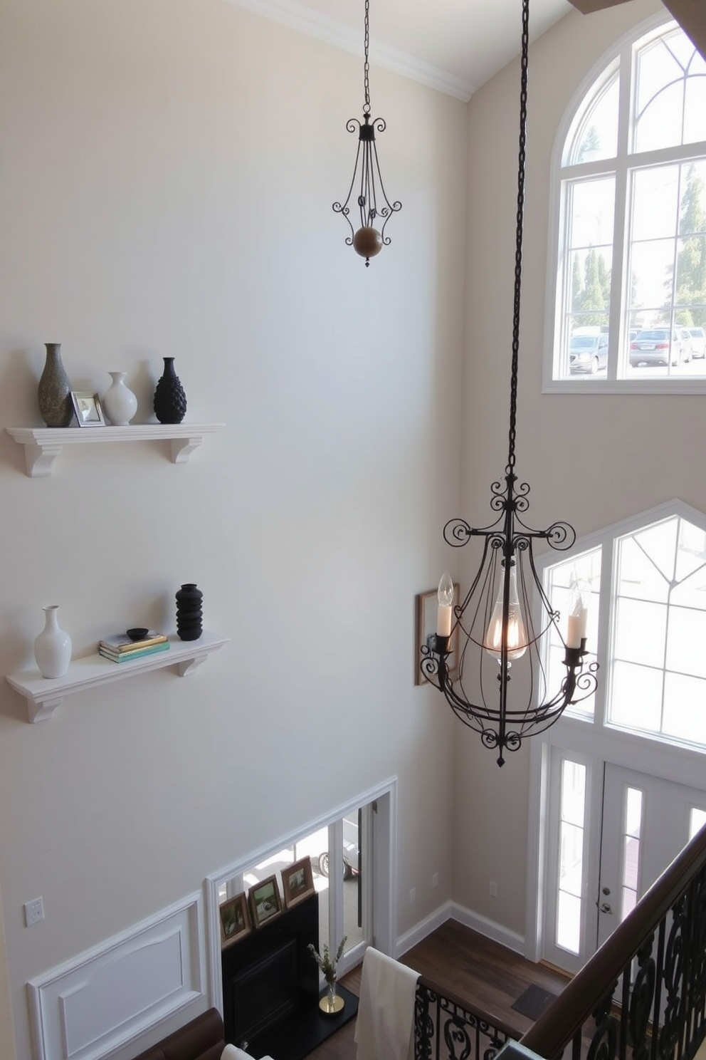 A pair of decorative ledges are installed on the walls, elegantly displaying an array of curated decor items such as vases and art pieces. The ledges are finished in a soft white color, complementing the surrounding wall paint and enhancing the overall aesthetic. The two story foyer features a grand staircase with intricate wrought iron railings, leading to a beautifully designed upper landing. Large windows allow natural light to flood the space, illuminating the elegant chandelier hanging from the high ceiling.