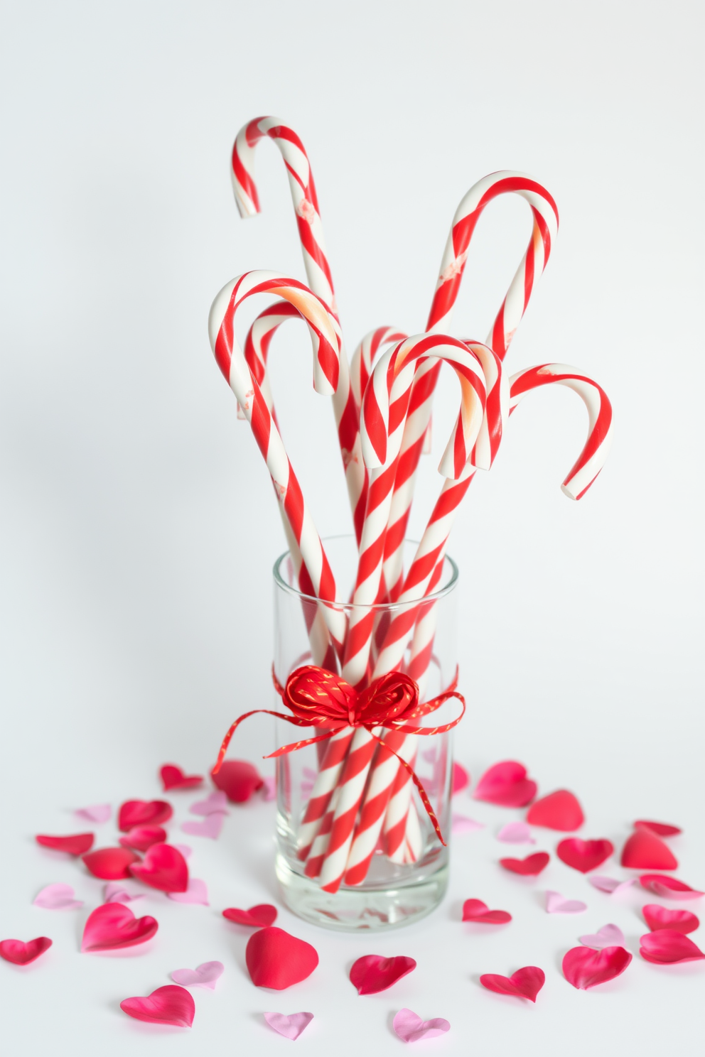 A charming Valentine's Day display featuring red and white striped candy canes arranged in a glass vase. Surrounding the vase are scattered rose petals and small heart-shaped decorations to enhance the romantic theme.