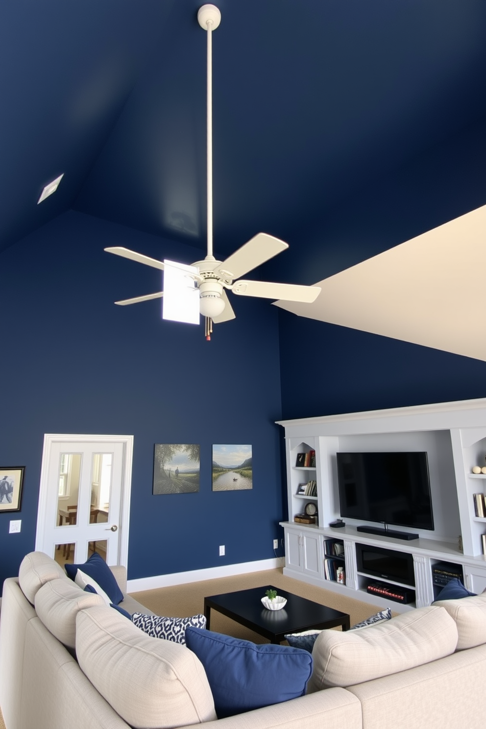 A bright white ceiling fan hangs from the high ceiling, providing a refreshing airflow throughout the space. The walls are painted in a deep navy blue, creating a cozy and inviting atmosphere for relaxation and entertainment. The room features a plush sectional sofa in a light gray fabric, perfect for lounging with friends. A large flat-screen TV is mounted on the wall opposite the sofa, surrounded by built-in shelves displaying books and collectibles.