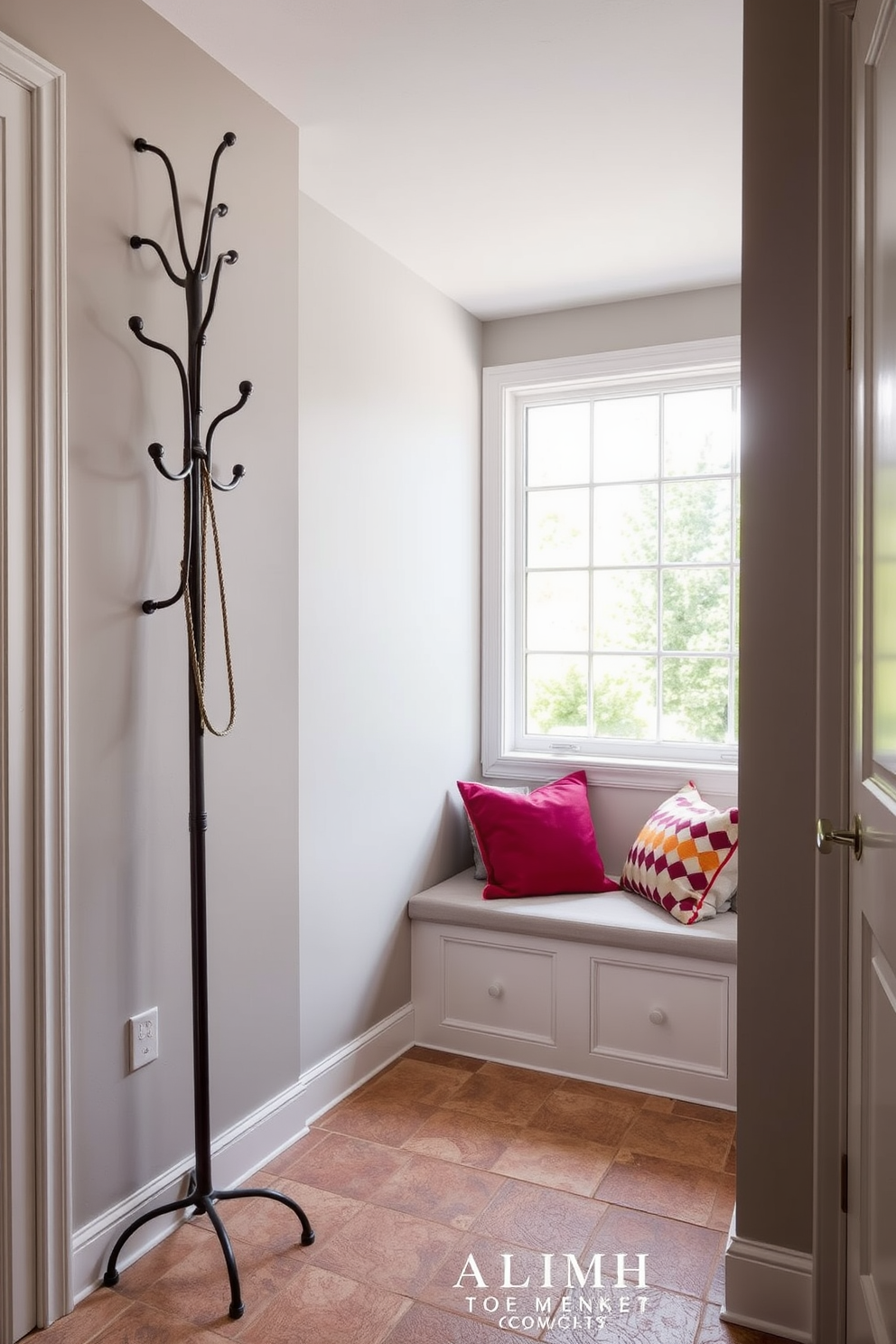 A stylish coat rack with artistic flair stands prominently in a well-organized mudroom. The walls are adorned with a soft gray hue, and the floor is covered with durable, textured tiles that add warmth to the space. The coat rack features unique hooks in varying shapes and sizes, providing both functionality and visual interest. Natural light floods the room through a large window, illuminating a cozy bench beneath it, accented with colorful cushions.