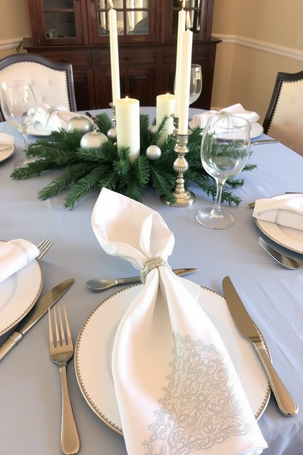 A beautifully set dining table for winter featuring elegant white and silver napkins folded in intricate designs. The table is adorned with a soft blue tablecloth, and a centerpiece of pine branches and white candles creates a warm, inviting atmosphere.