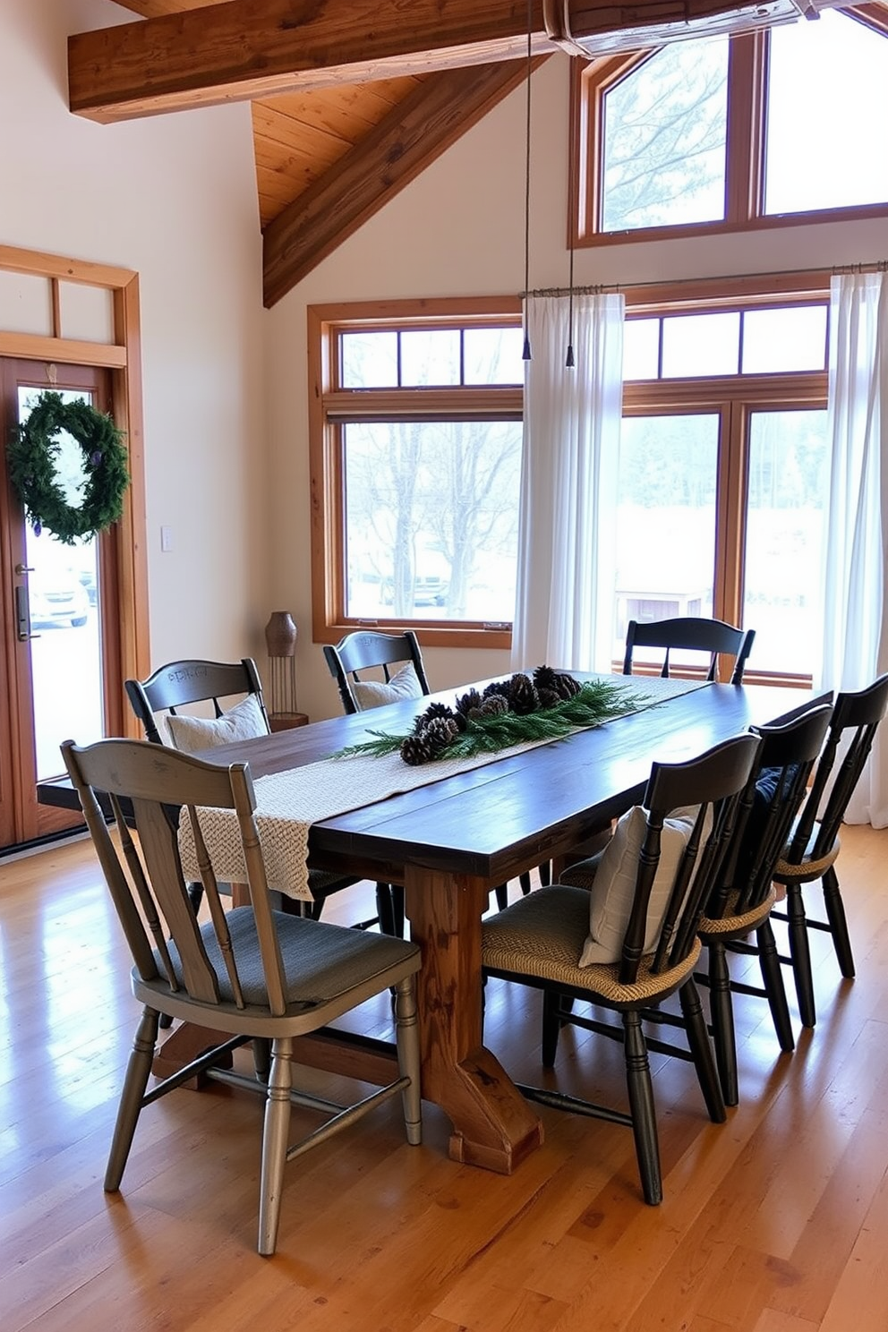 A cozy winter dining room features a large rustic wooden table surrounded by mismatched chairs, each with soft, warm cushions. The table is adorned with a centerpiece of pinecones and evergreen branches, while a knitted table runner adds texture and warmth to the setting. The walls are painted in a warm cream color, complemented by wooden beams that enhance the rustic charm. Large windows allow natural light to flood the room, with sheer white curtains gently framing the views of a snowy landscape outside.