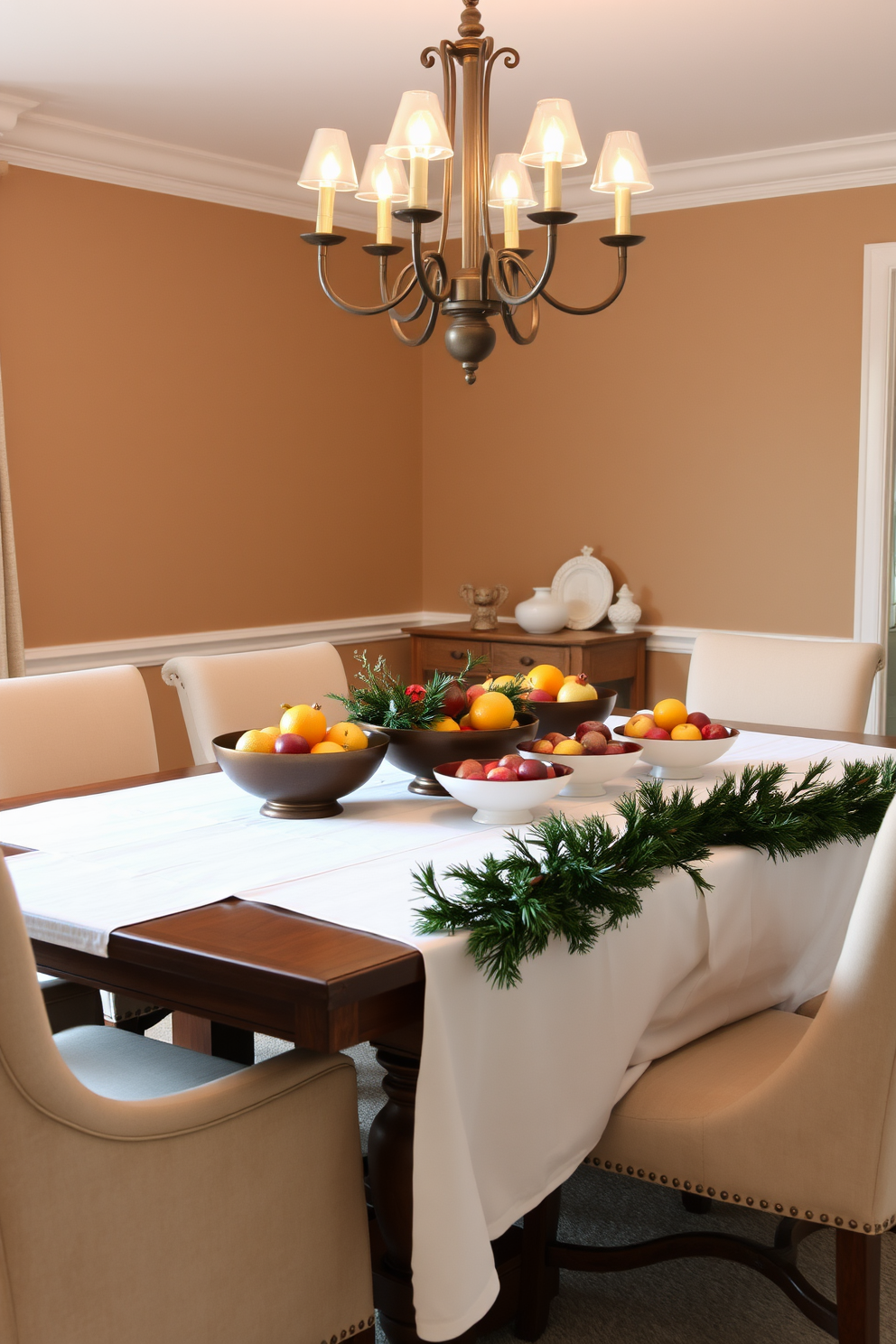 A cozy winter dining room setting featuring a large wooden dining table adorned with a crisp white tablecloth. On the table, elegant bowls filled with seasonal fruits like oranges, pomegranates, and apples create a vibrant centerpiece. The walls are painted in a warm taupe hue, complemented by soft, ambient lighting from a stylish chandelier above. Plush upholstered chairs surround the table, and a festive garland drapes across the table's edge, adding a touch of seasonal charm.