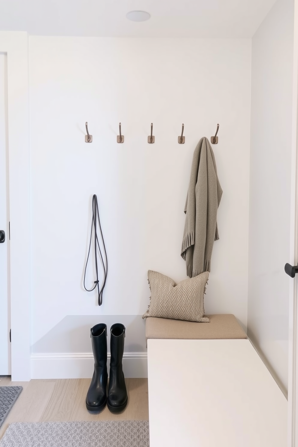 A modern winter mudroom features sleek metal hooks mounted on a smooth white wall, providing a minimalist yet functional touch. Beneath the hooks, a stylish bench with a cozy cushion invites guests to sit while removing their boots, creating a warm and welcoming entryway.