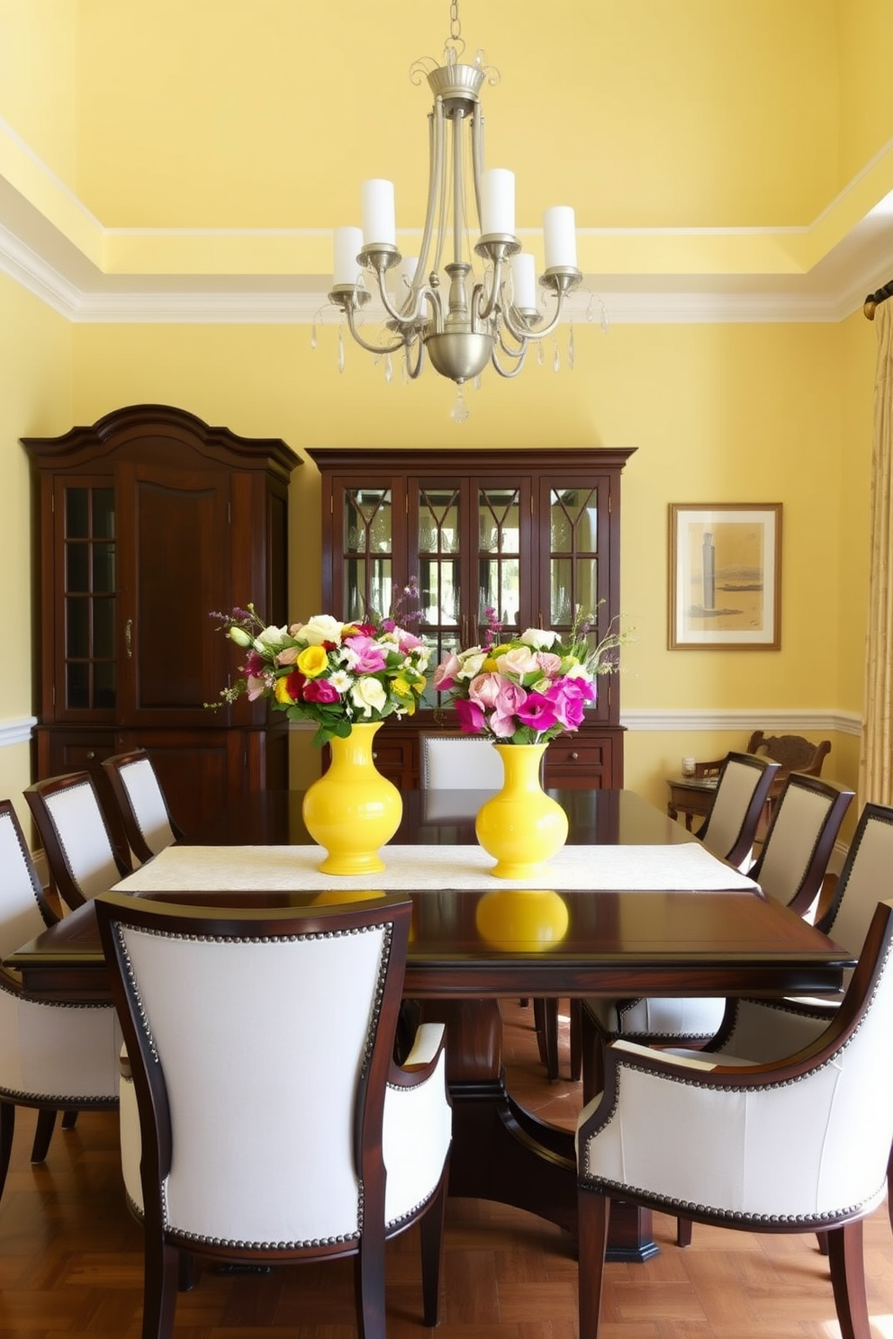 A bright and inviting dining room features a large wooden table surrounded by elegant upholstered chairs. The walls are painted in a soft yellow hue, creating a warm and cheerful atmosphere. On the table, two yellow vases filled with fresh floral displays add a vibrant touch to the decor. A stylish chandelier hangs above, providing a focal point and enhancing the overall ambiance of the space.