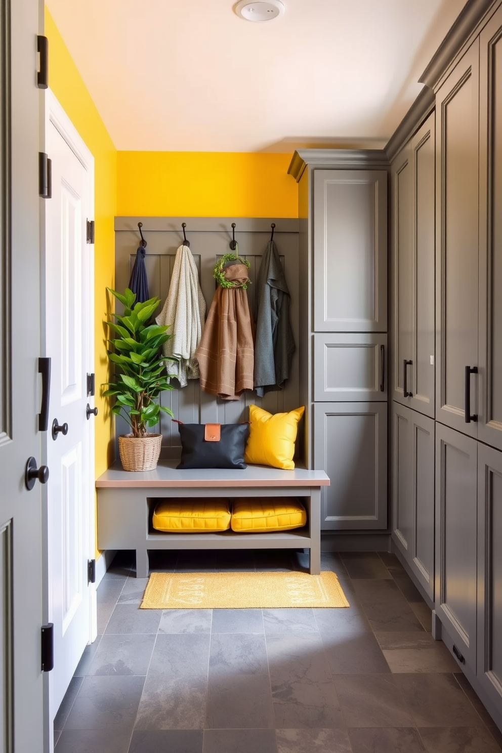 A welcoming mudroom features a yellow accent wall paired with gray cabinetry. The floor is covered with a durable gray tile, and a stylish bench with yellow cushions provides seating. Hooks for coats are mounted on the wall above the bench, complemented by a decorative yellow rug underneath. Potted plants add a touch of greenery, enhancing the warmth of the space.